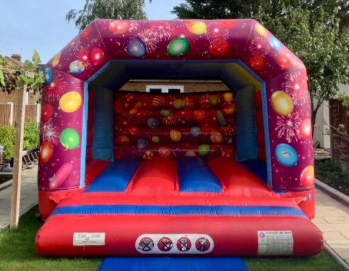 Vibrant red and blue inflatable bouncy castle with balloon and firework designs set up outdoors, provided by Bizi Bouncers UK for Children's Party Planner Wanstead.