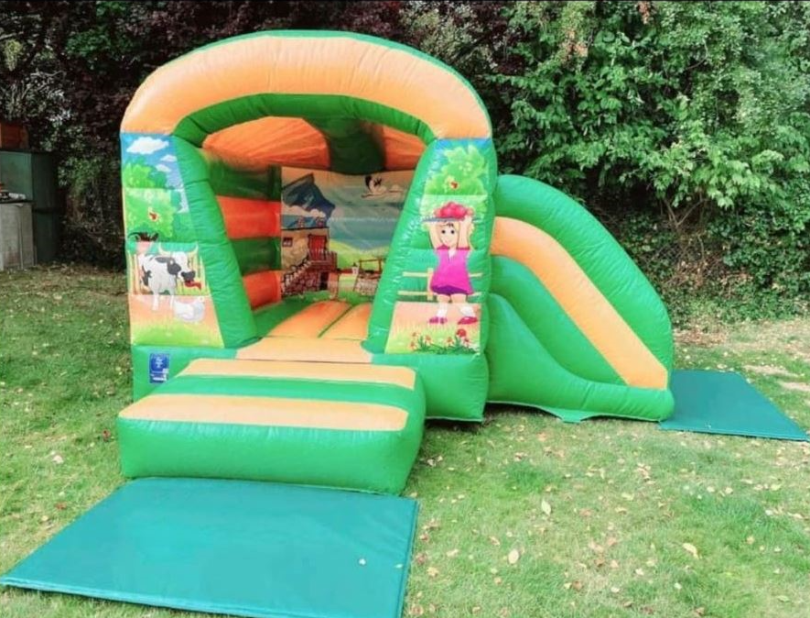 Farmhouse-themed inflatable bouncy castle with slide for kids by Bizi Bouncers UK, set up outdoors on grass