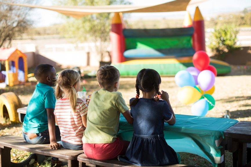 Children sitting at a party table with balloons and a bouncy castle in the background, provided by Bizi Bouncers UK for Party Equipment Rental Service in Barkingside